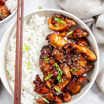 two bowls with chicken and rice inside them. gray towel in the back along with chopsticks and sesame seeds