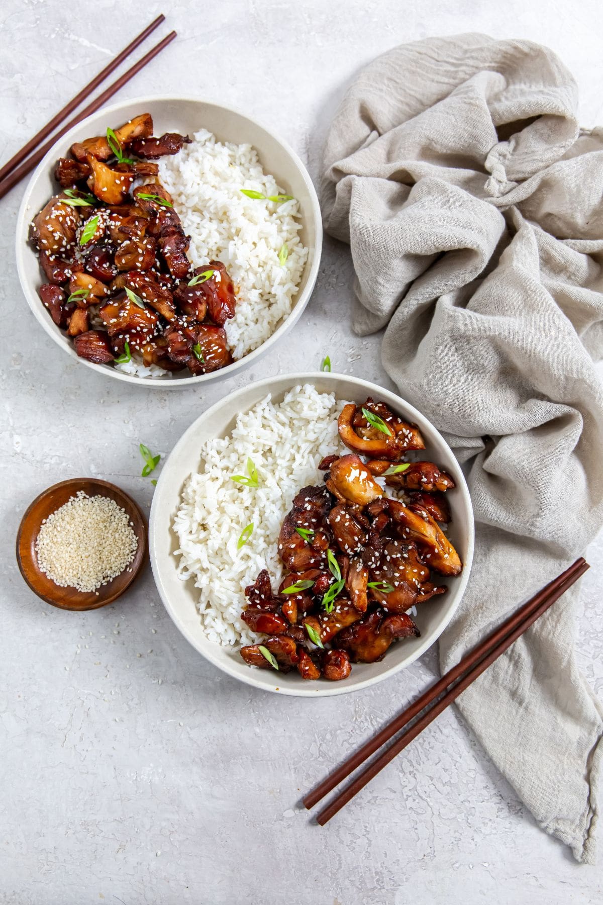 two bowls with chicken and rice inside them. gray towel in the back along with chopsticks and sesame seeds