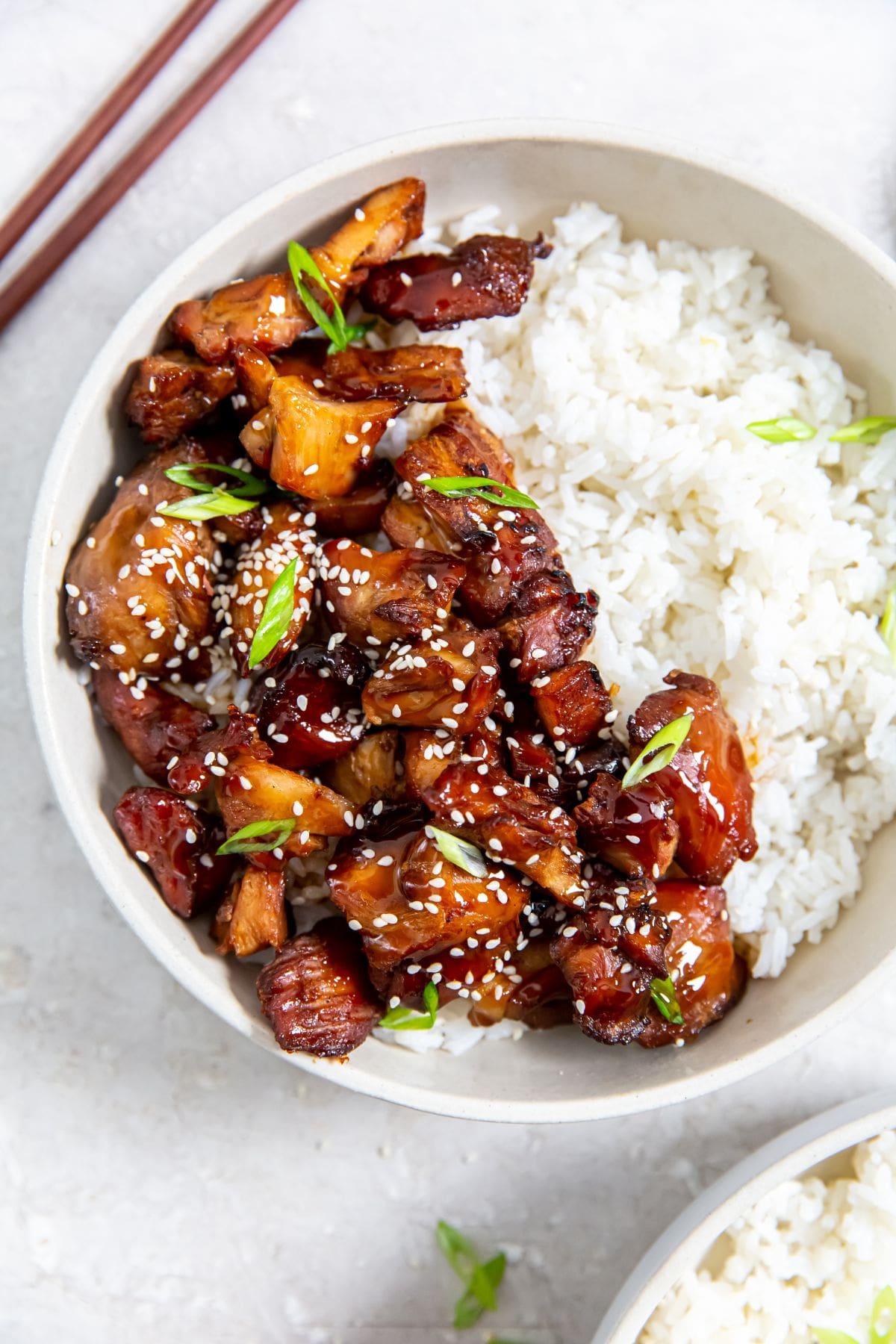 two bowls with chicken and rice inside them. gray towel in the back along with chopsticks and sesame seeds