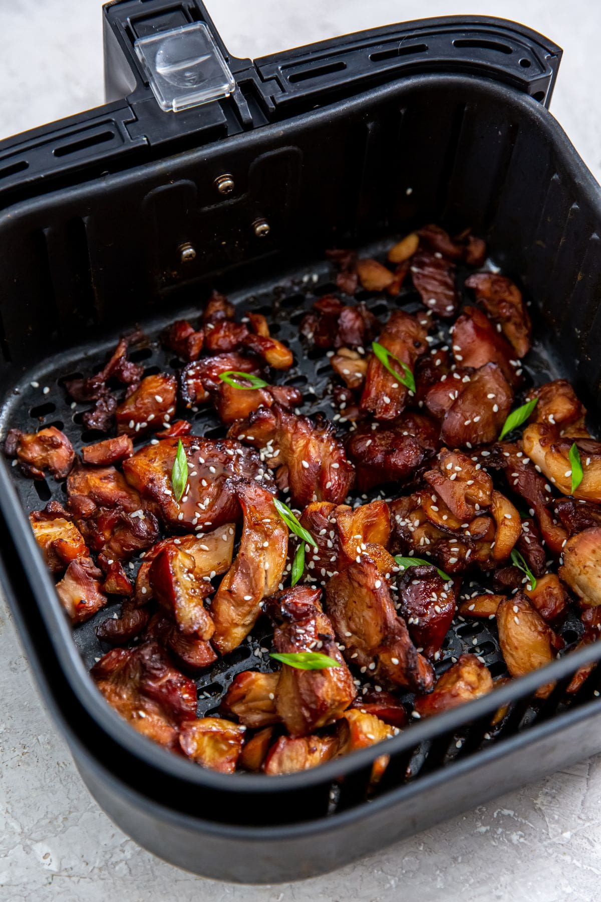 black air fryer basket with cooked chicken inside. tossed in sauce scallions and sesame seeds 