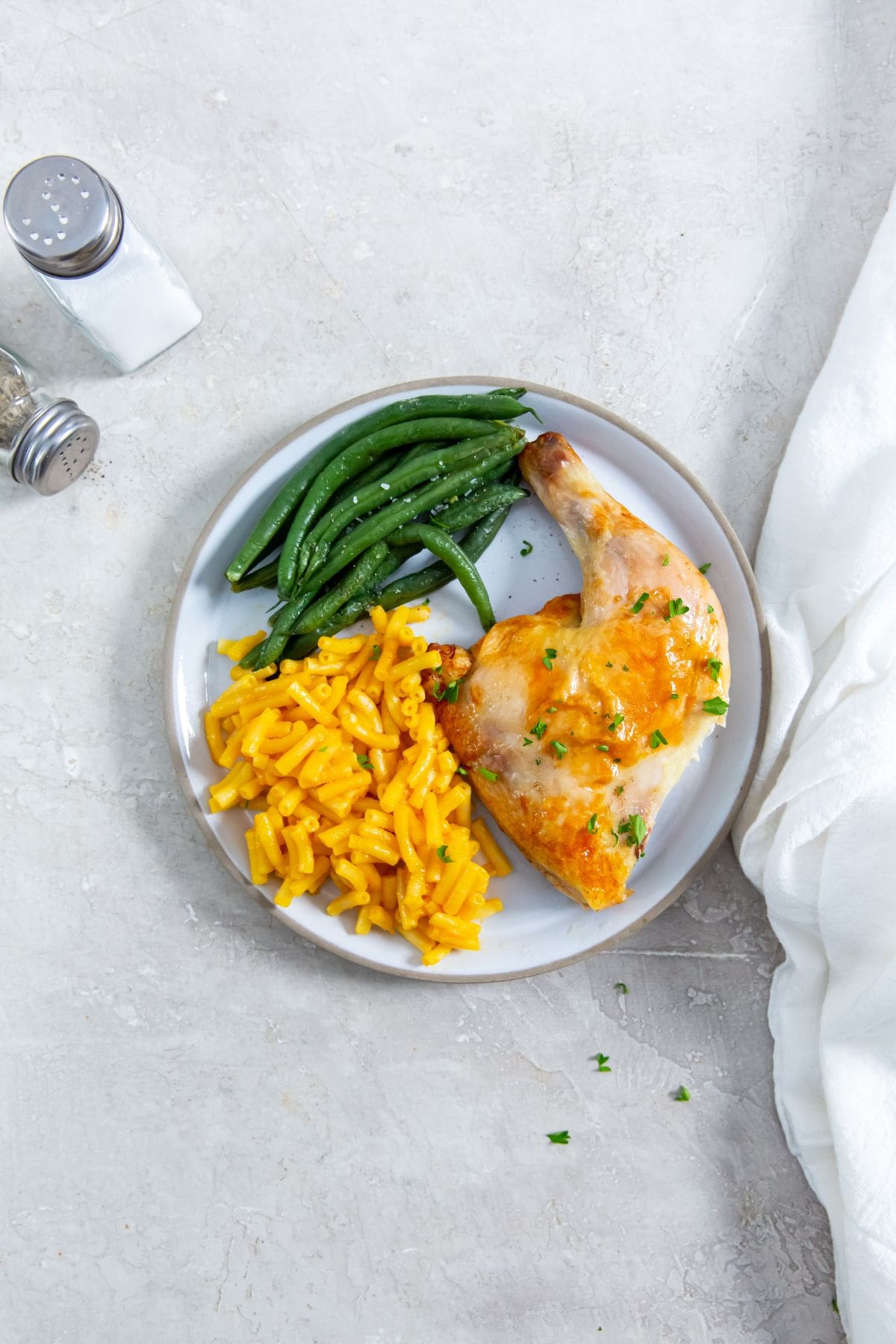 White plate with chicken Mac and cheese, and green beans on it. white towel in the back with salt and black pepper shaker