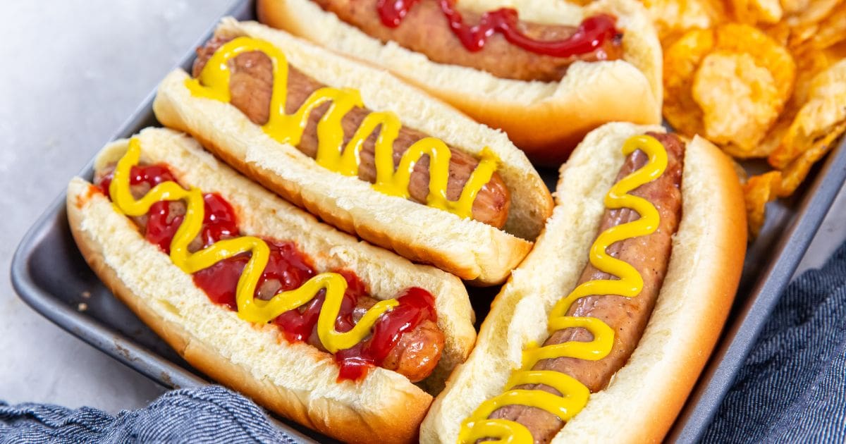 tray with chicken in a hotdog bun mustard and ketchup on top. chips on the side. blue towel behind it.