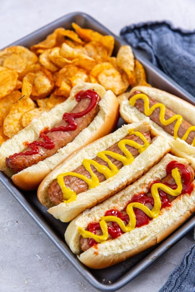 tray with chicken in a hotdog bun mustard and ketchup on top. chips on the side. blue towel behind it.