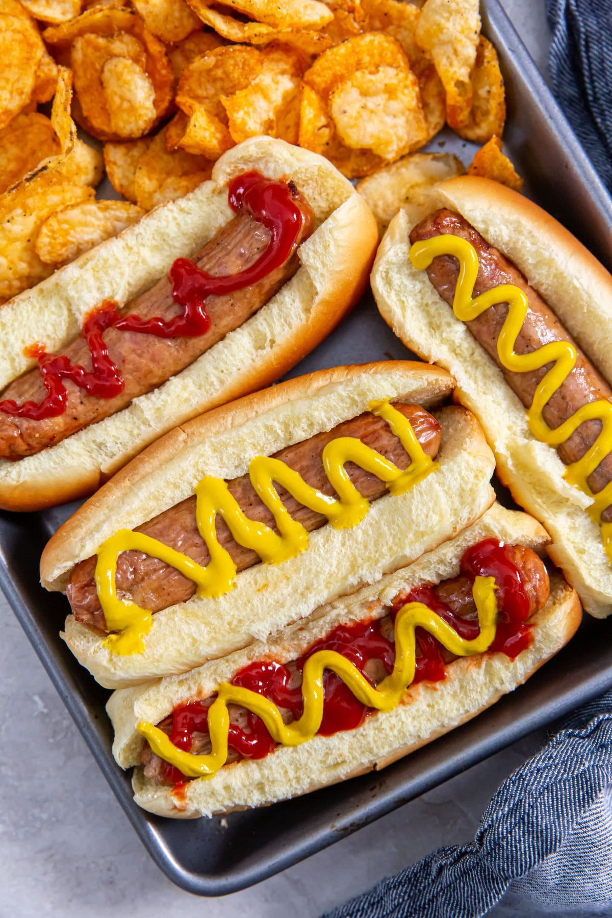 tray with chicken in a hotdog bun mustard and ketchup on top. chips on the side. blue towel behind it.