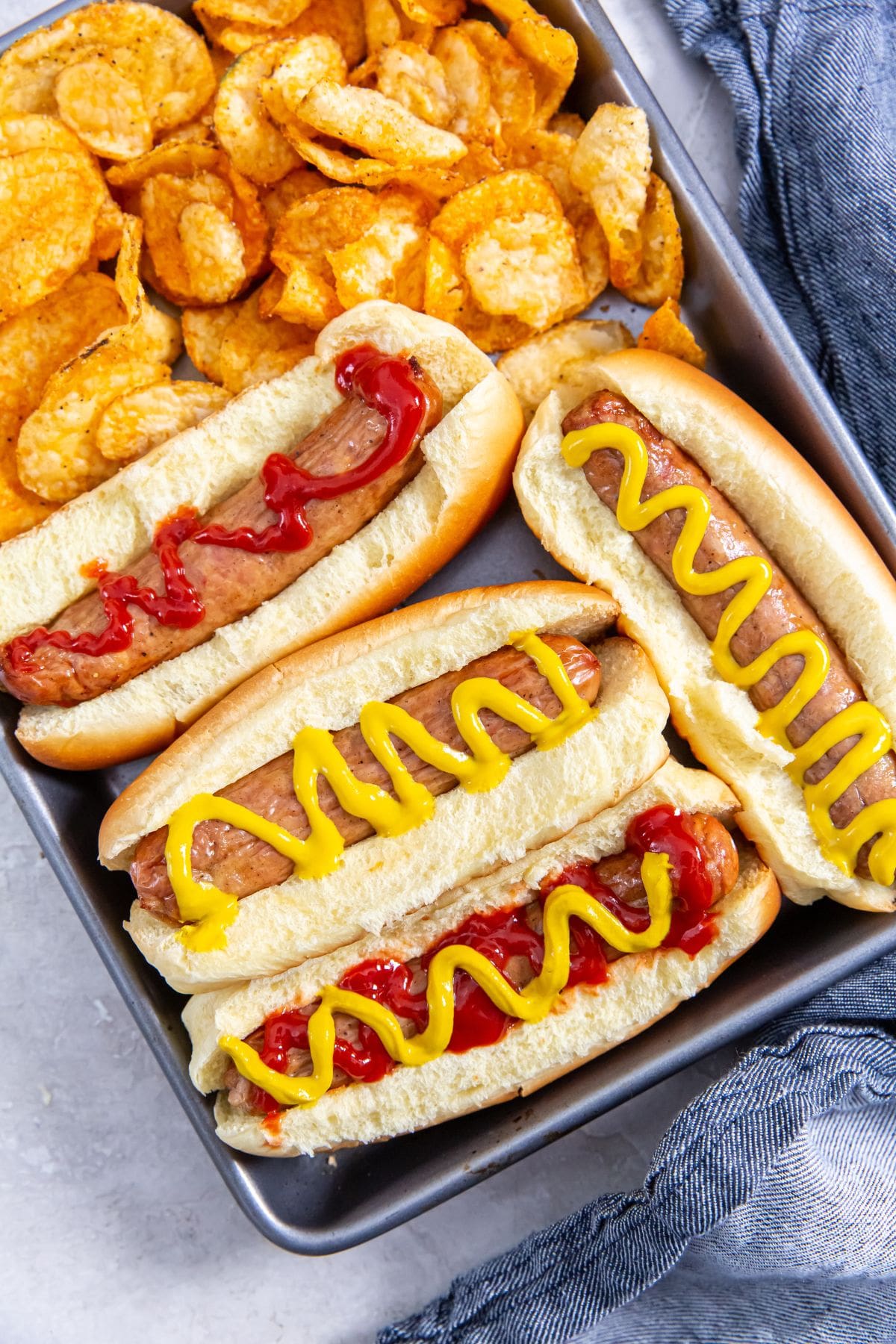 tray with chicken in a hotdog bun mustard and ketchup on top. chips on the side. blue towel behind it.