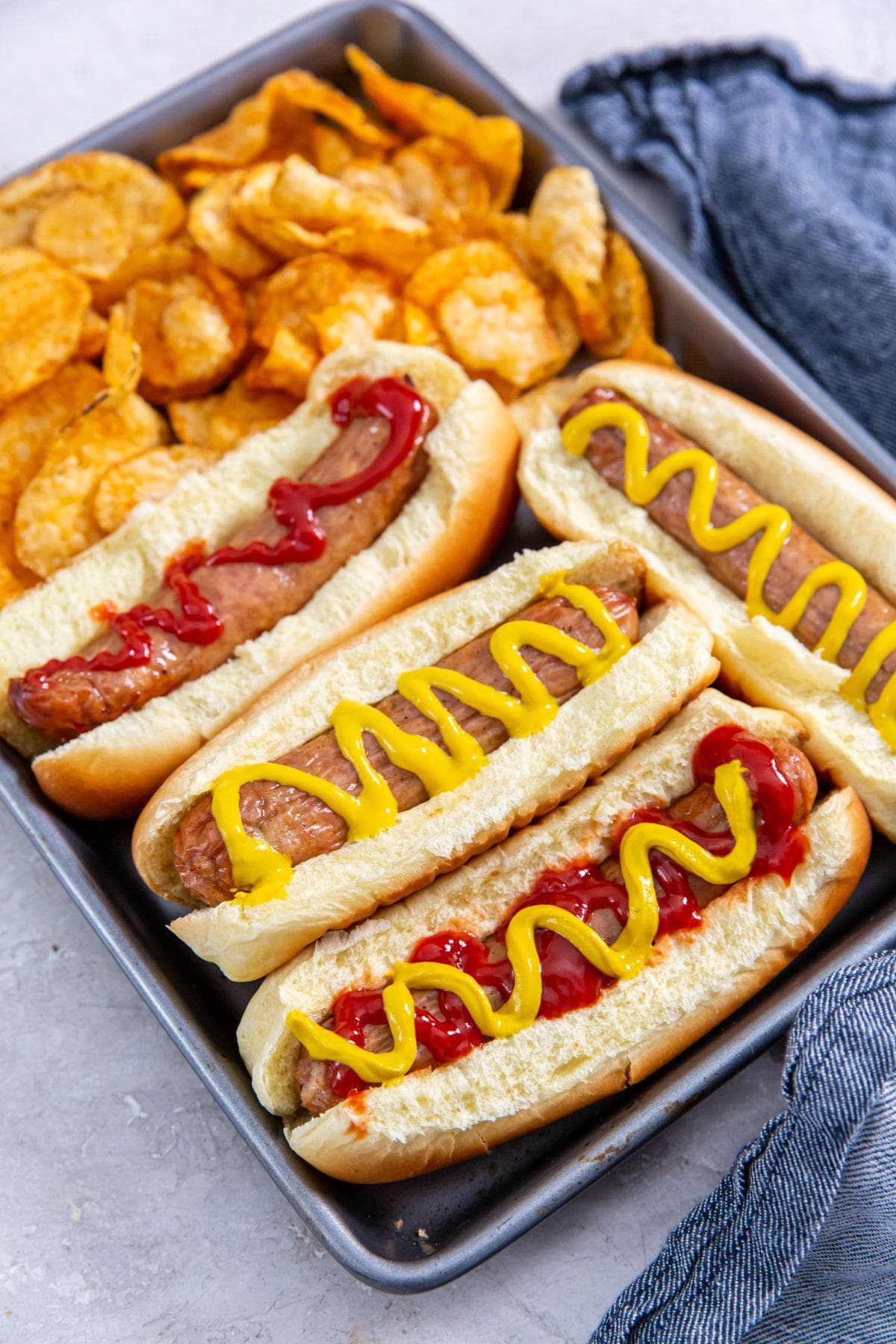 tray with chicken in a hotdog bun mustard and ketchup on top. chips on the side. blue towel behind it.