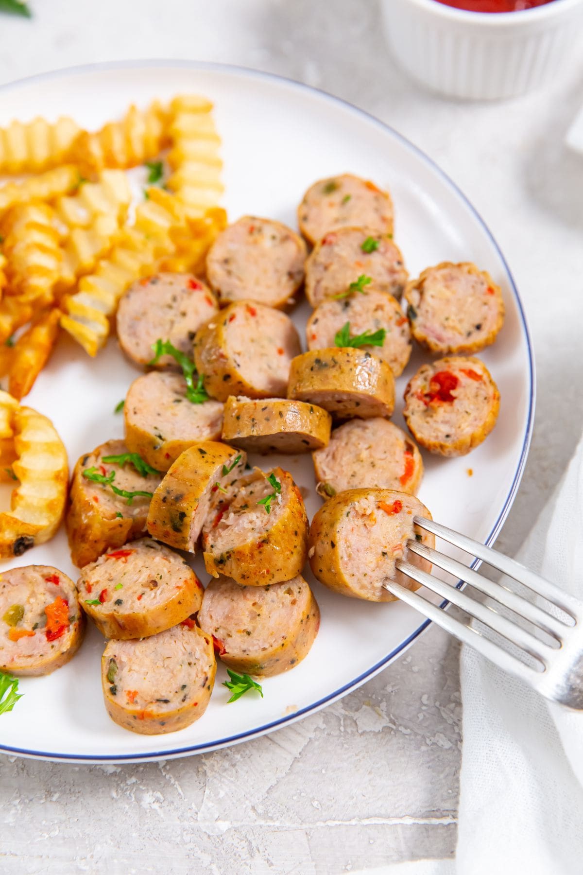 white plate with chicken sausage and fries on the plate. parsley, ketchup and white towel in the background