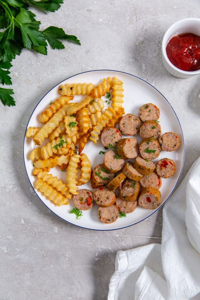 white plate with chicken sausage and fries on the plate. parsley, ketchup and white towel in the background