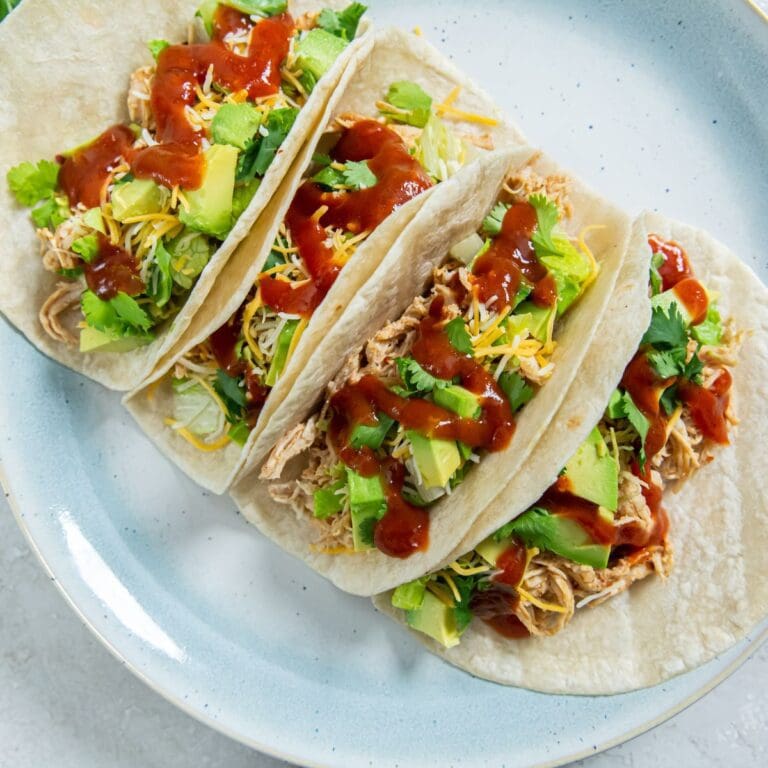 Blue plate with four tacos on it. shredding chicken and avocado in the back