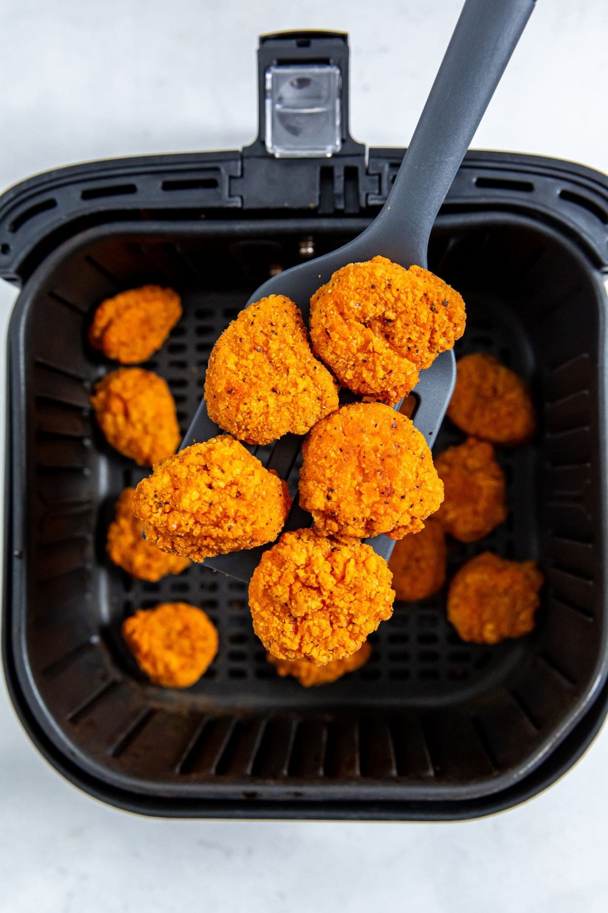 black air fryer basket with cooked chicken inside. spatula holding some nuggets.