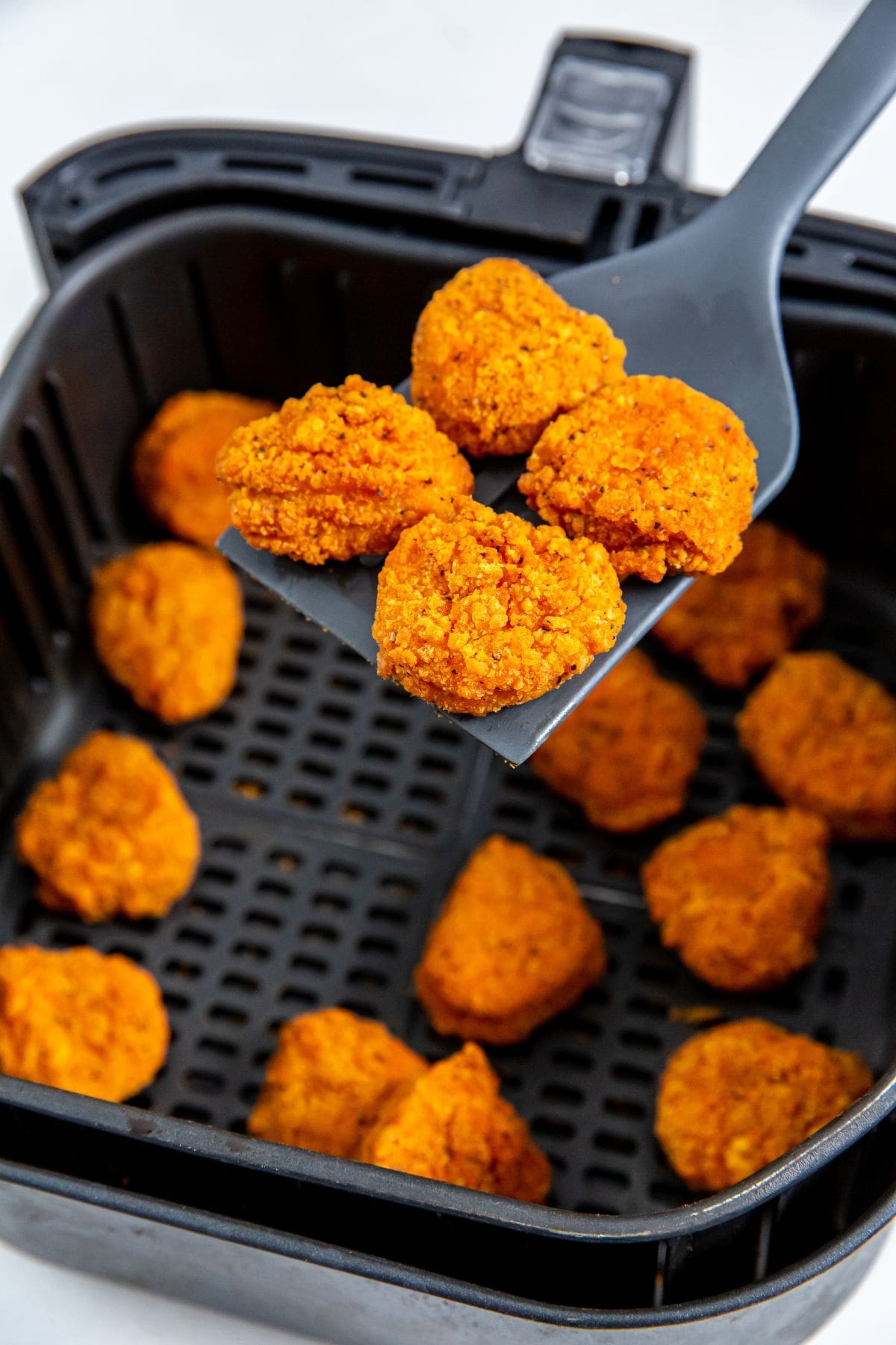 black air fryer basket with cooked chicken inside. spatula holding some nuggets. 