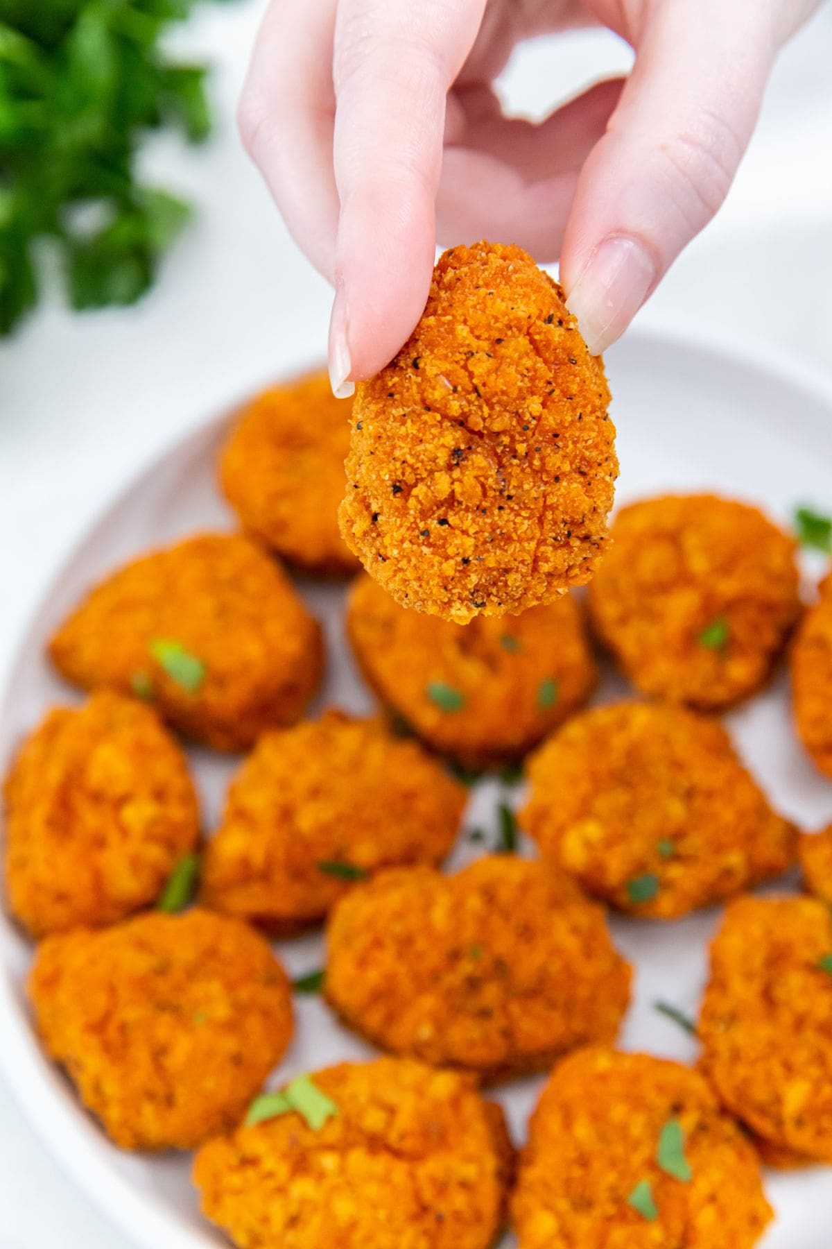 white plate of chicken nuggets. one nugget getting held above the plate. parsley in the back