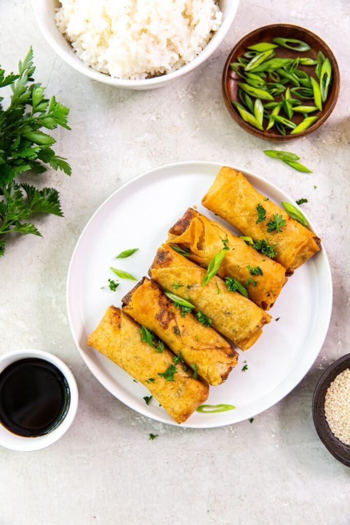 white plate with spring rolls on it. topped with parsley. Scallions, white rice, soy sauce, and sesame seeds.