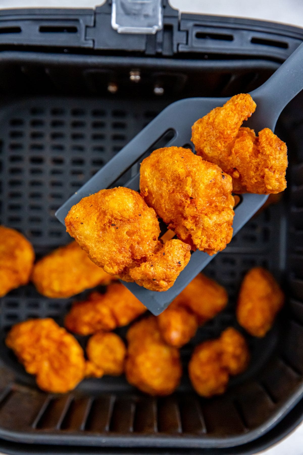 Black air fryer with cooked chicken inside. spatula holding some chicken up.