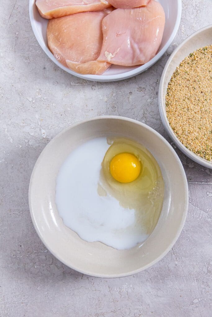 three bowls egg wash, bread crumbs, and chicken cutlets