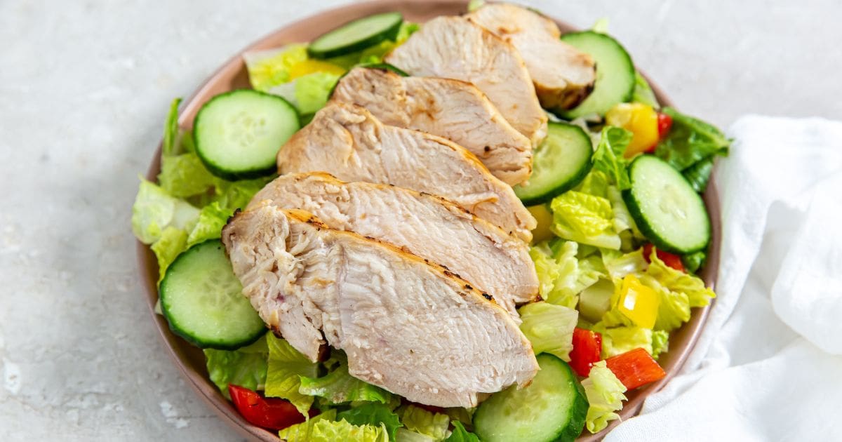 chicken on the top of a salad with cucumbers and tomatoes on a plate.