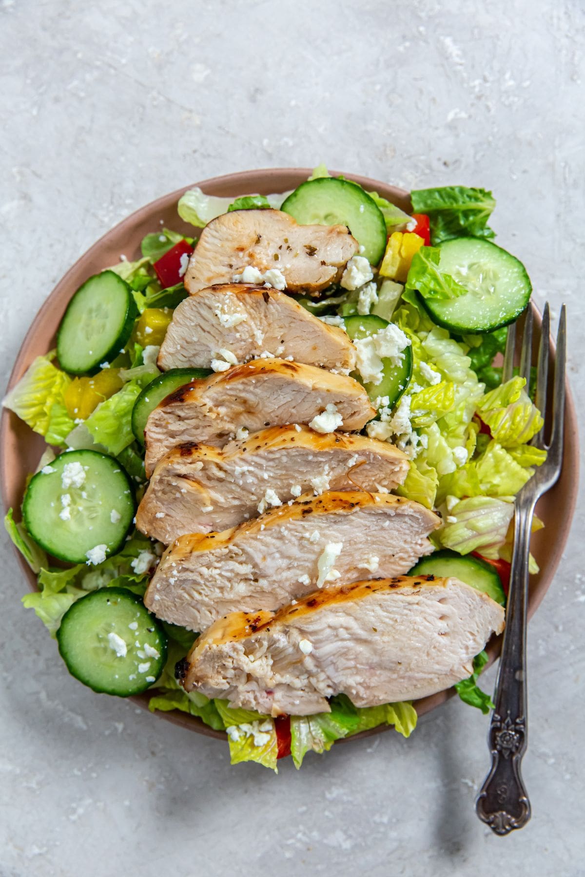 chicken on the top of a salad with cucumbers and tomatoes on a plate.