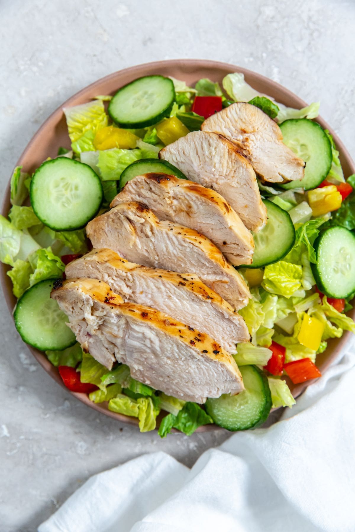 chicken on the top of a salad with cucumbers and tomatoes on a plate.