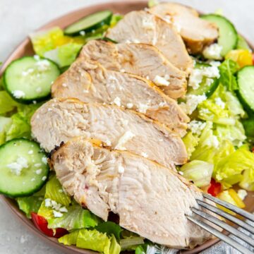 chicken on the top of a salad with cucumbers and tomatoes on a plate.