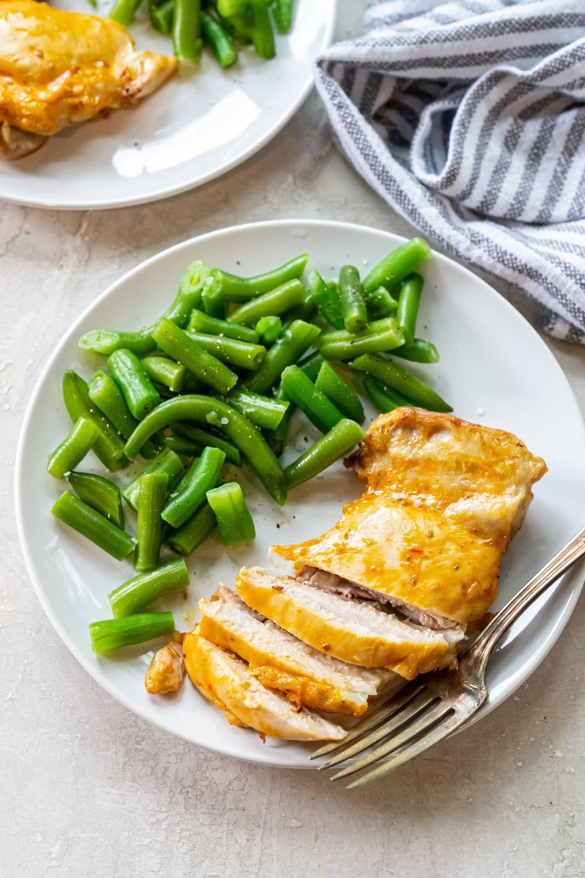 white plate with chicken and green beans on it. fork next to chicken. striped towel next to plates.