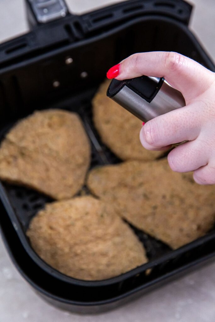 Air Fryer Chicken Cutlets black air fryer basket with raw chicken inside. Person spraying chicken over the top.