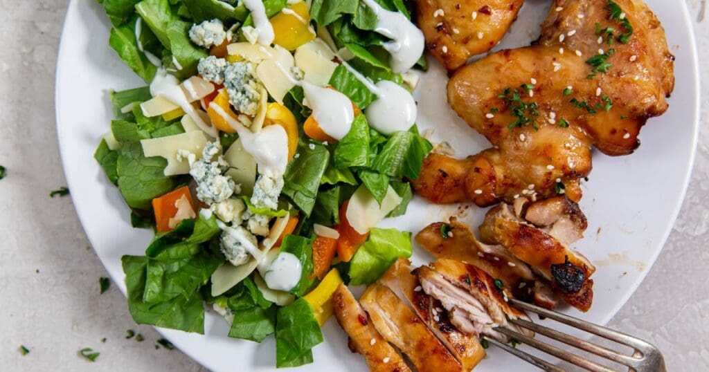 A plate of grilled chicken thighs with a side salad of lettuce, bell peppers, carrots, and blue cheese dressing.