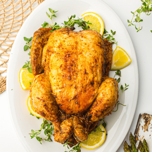 Air Fryer whole chicken on a white plate garnished with lemon slices and fresh herbs, served on a table with asparagus on the side.