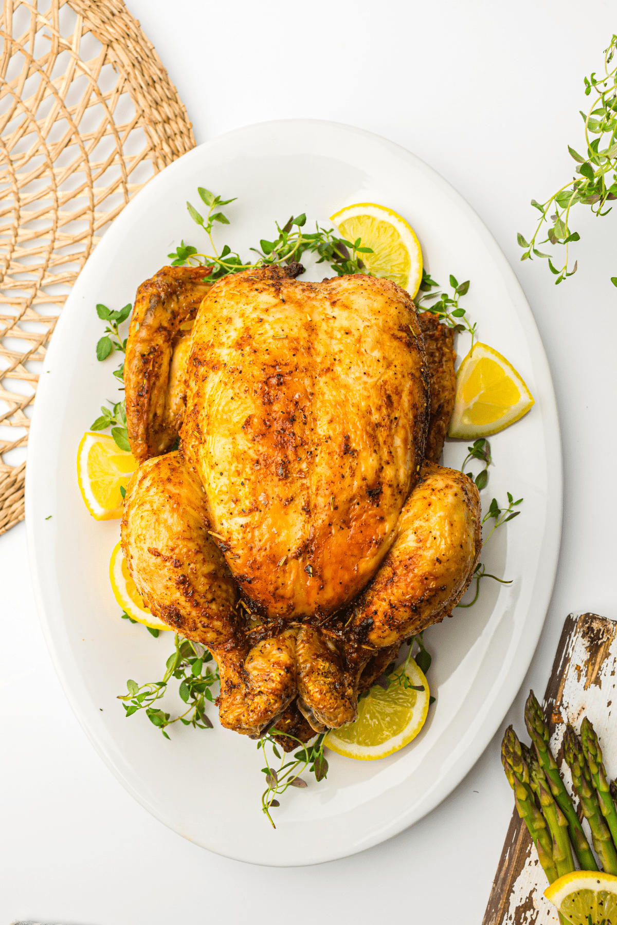 Air Fryer whole chicken on a white plate garnished with lemon slices and fresh herbs, served on a table with asparagus on the side.