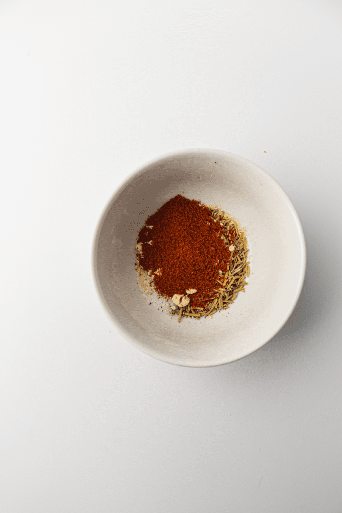 A white bowl containing a mix of red chili powder and green dried herbs, perfect for seasoning an air fryer whole chicken, on a white surface.