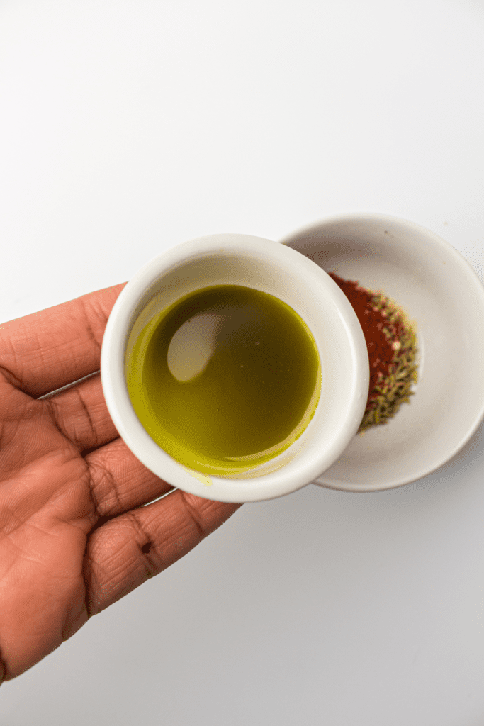 A person holds a small bowl of olive oil in their hand above a larger bowl containing spices for an air fryer whole chicken recipe.