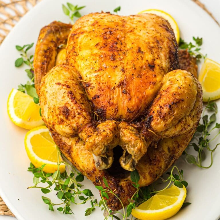 An air fryer roasted chicken garnished with lemon slices and fresh herbs on a white plate.