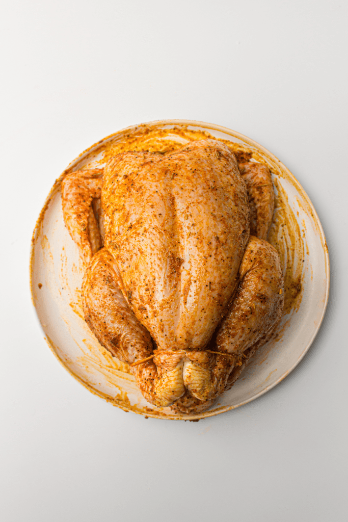 Whole raw chicken seasoned with spices on a white plate.