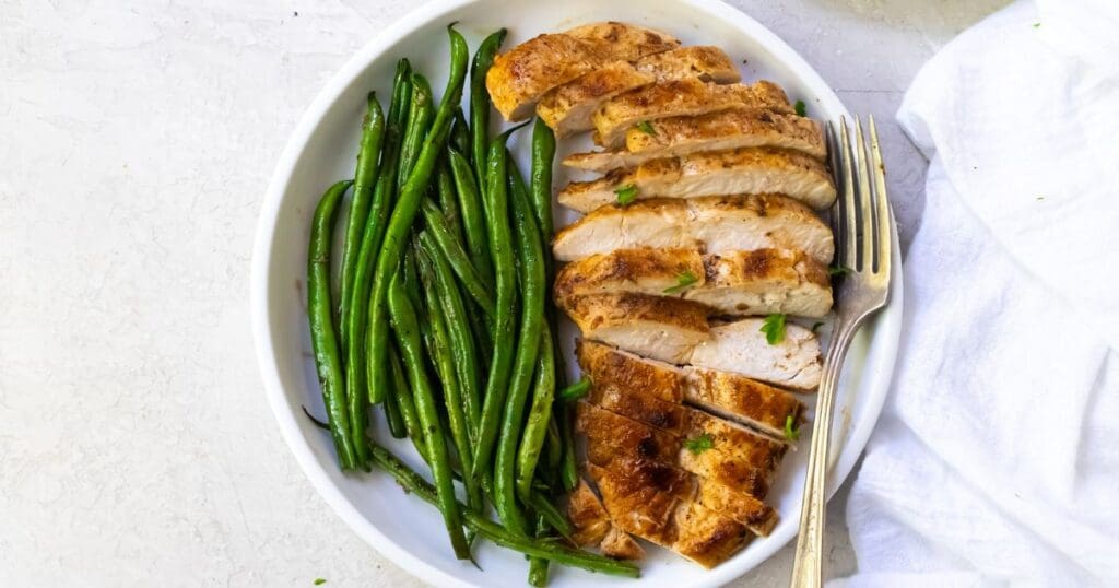 Sliced grilled chicken breast and green beans on a white oval dish, served on a marble surface with a white cloth on the side.