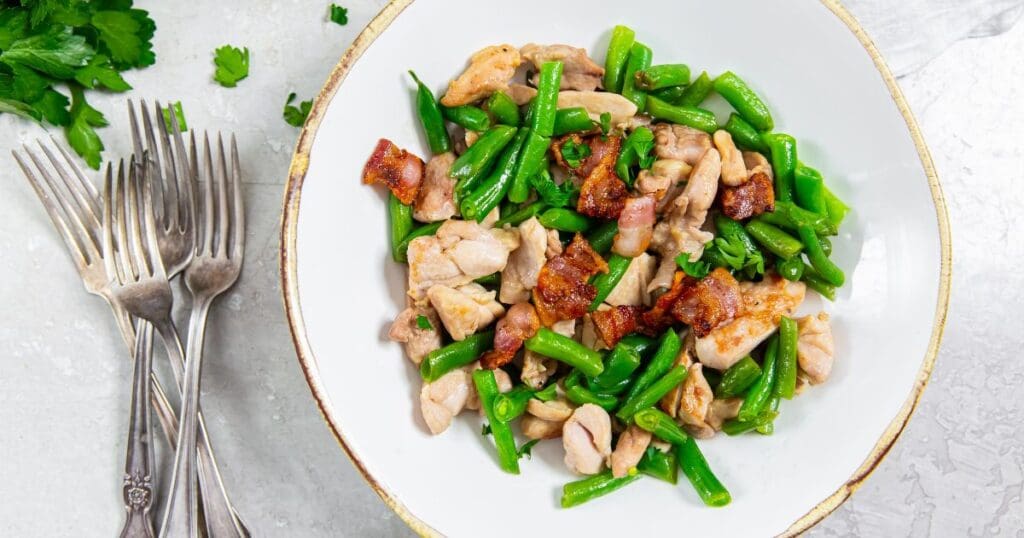 Plate of stir-fried chicken and green beans with bacon, served with a fork and spoon, on a white and gold rimmed plate.