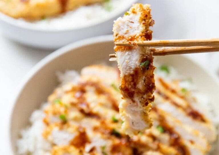 Close-up of chopsticks holding a piece of crispy, breaded chicken above a bowl of rice topped with more pieces of chicken and garnished with green onions. Discover tasty Chicken Air Fryer Recipes that make it easier than ever to get that perfect crunch.