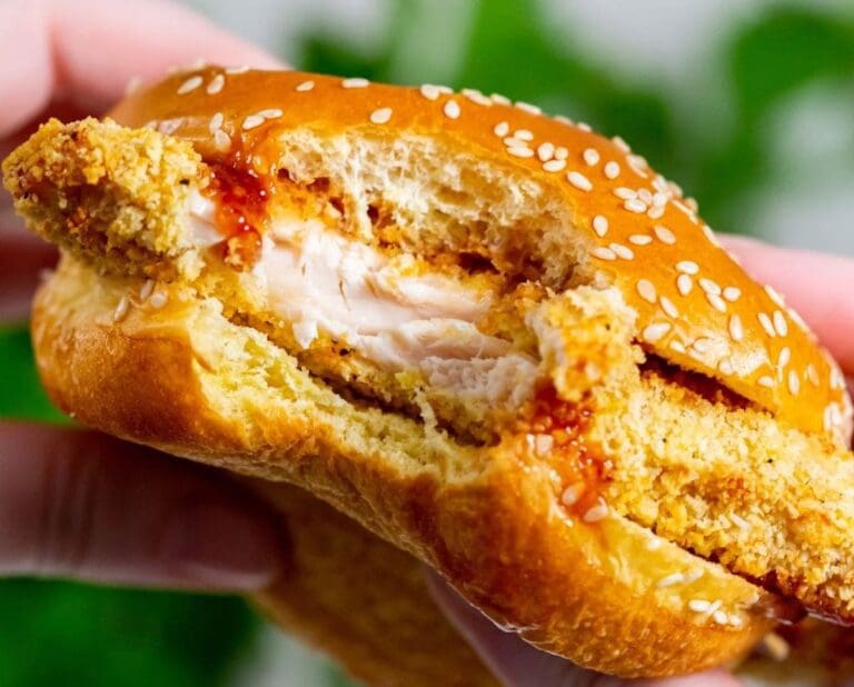 Close-up of a partially eaten sesame seed bun sandwich filled with a crispy chicken patty and some sauce, held by a hand. Perfect for those seeking tasty recipes using their air fryer.