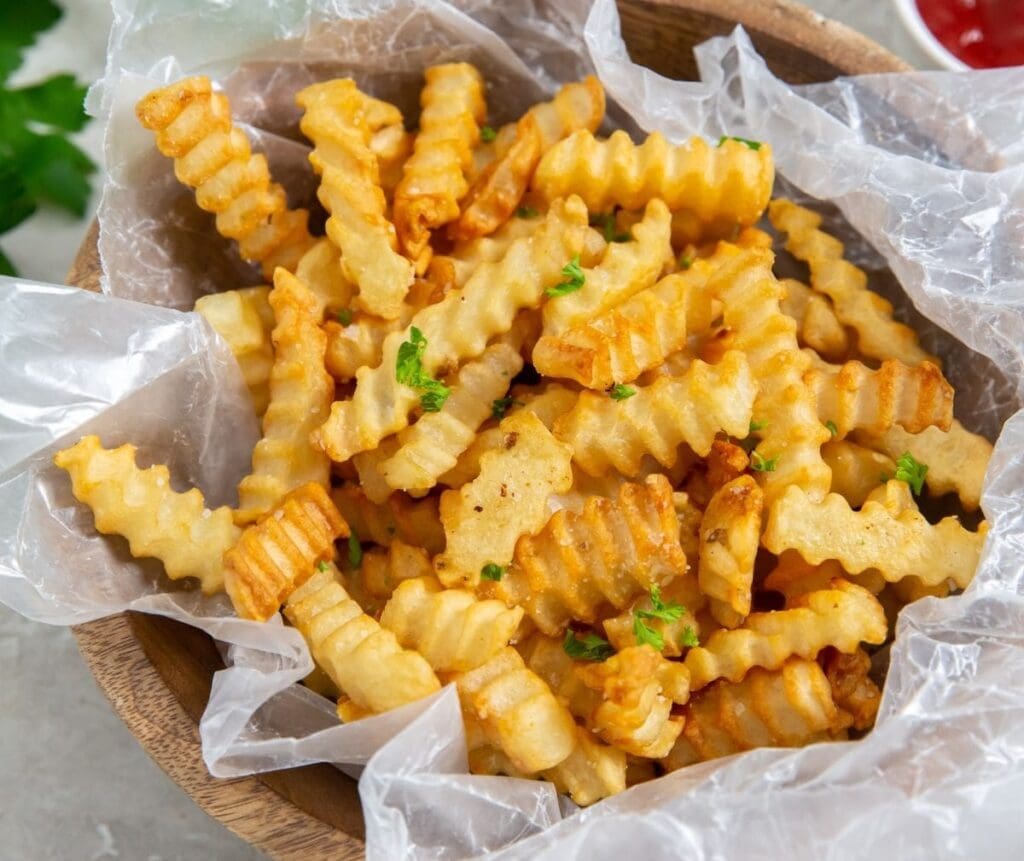 A wooden bowl filled with crinkle-cut fries garnished with green herbs, lined with translucent paper—mouthwatering sides to complement your mains.