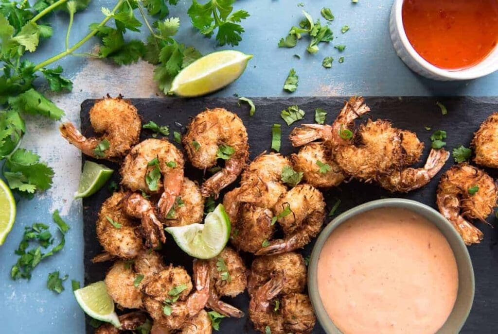 A platter of coconut shrimp garnished with lime wedges, accompanied by a bowl of pink dipping sauce and a separate cup of red sauce, with scattered cilantro on a blue surface—a perfect air fryer recipe ready in a flash for those busy nights.