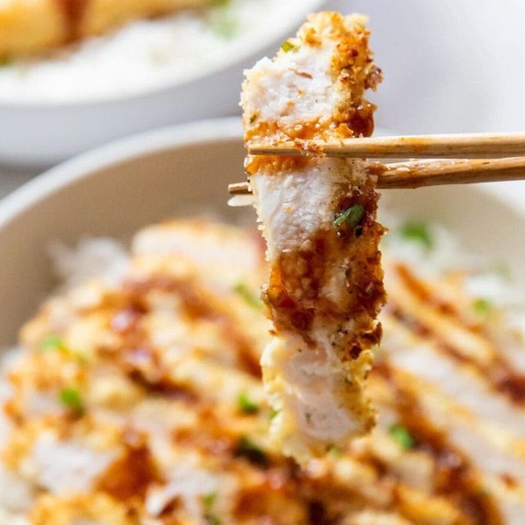 20 Easy Dinners My Family Claims Are Restaurant-Quality Close Up of a Piece of Katsu Chicken in Chopsticks Above a Bowl of Katsu Chicken and Rice