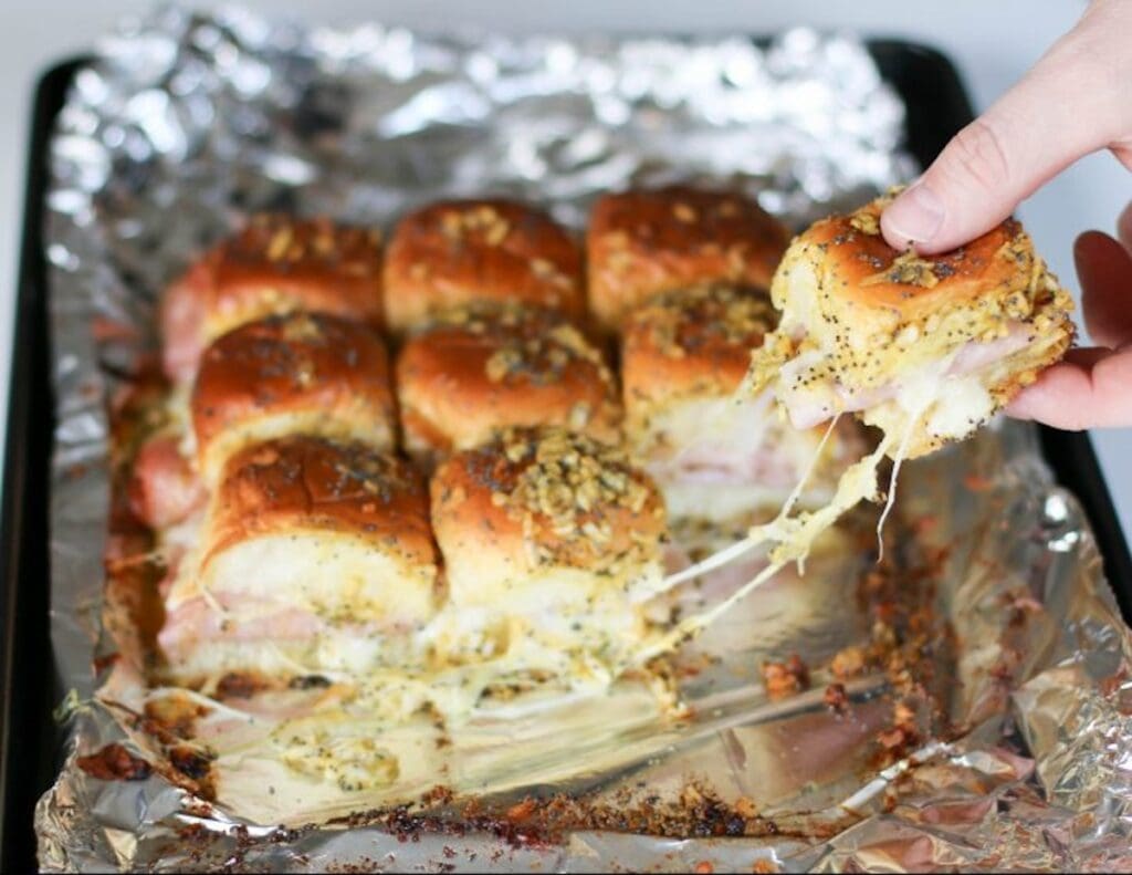 A hand is pulling a piece of a baked cheese and ham slider from a batch on a foil-covered tray. The melted cheese stretches between the piece and the remaining sliders, making it one of those must-try appetizers for any gathering.