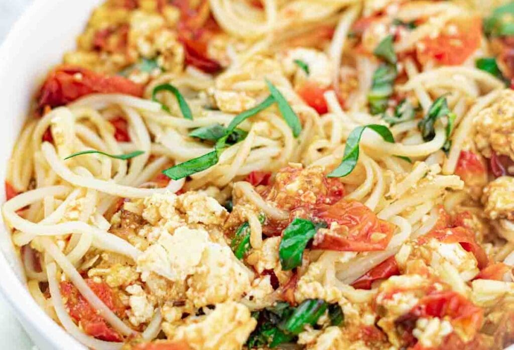 A close-up of a plate of spaghetti mixed with diced tomatoes, crumbled cheese, and garnished with fresh basil is perfect for those looking to whip up tasty dinners effortlessly.