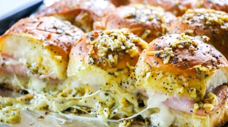 Close-up of baked sliders with melted cheese, ham, and a topping of chopped onions, poppy seeds, and seasoning on a baking tray. Perfect appetizers for any gathering!