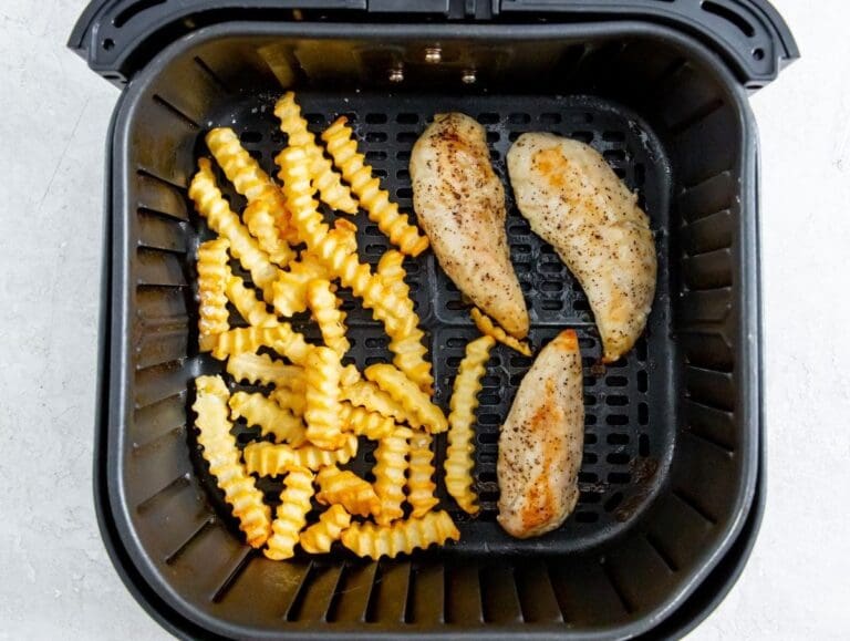 A black air fryer basket containing delicious crinkle-cut fries on the left side and seasoned chicken tenders on the right side. Discover this mouth-watering combo in our Air Fryer Recipes this month!