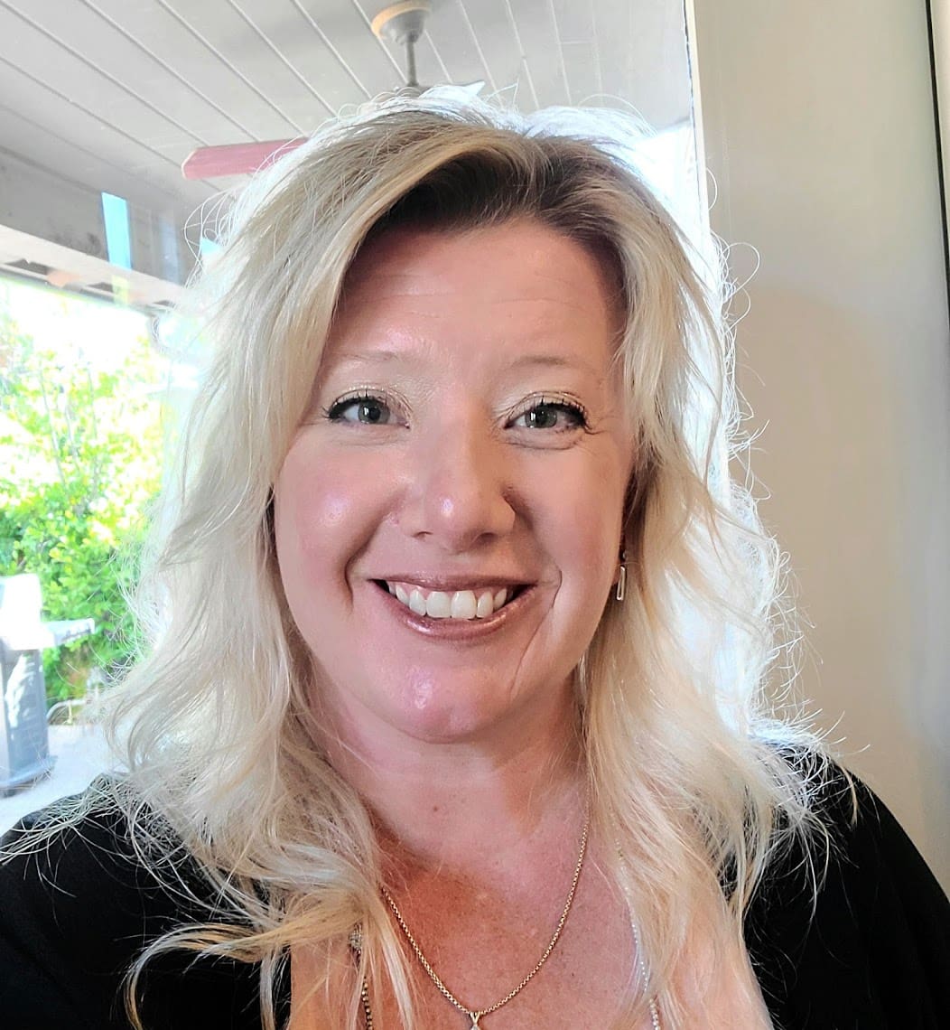 A person with long blond hair and a black top is smiling indoors near a window, enjoying the greenery outside their home.