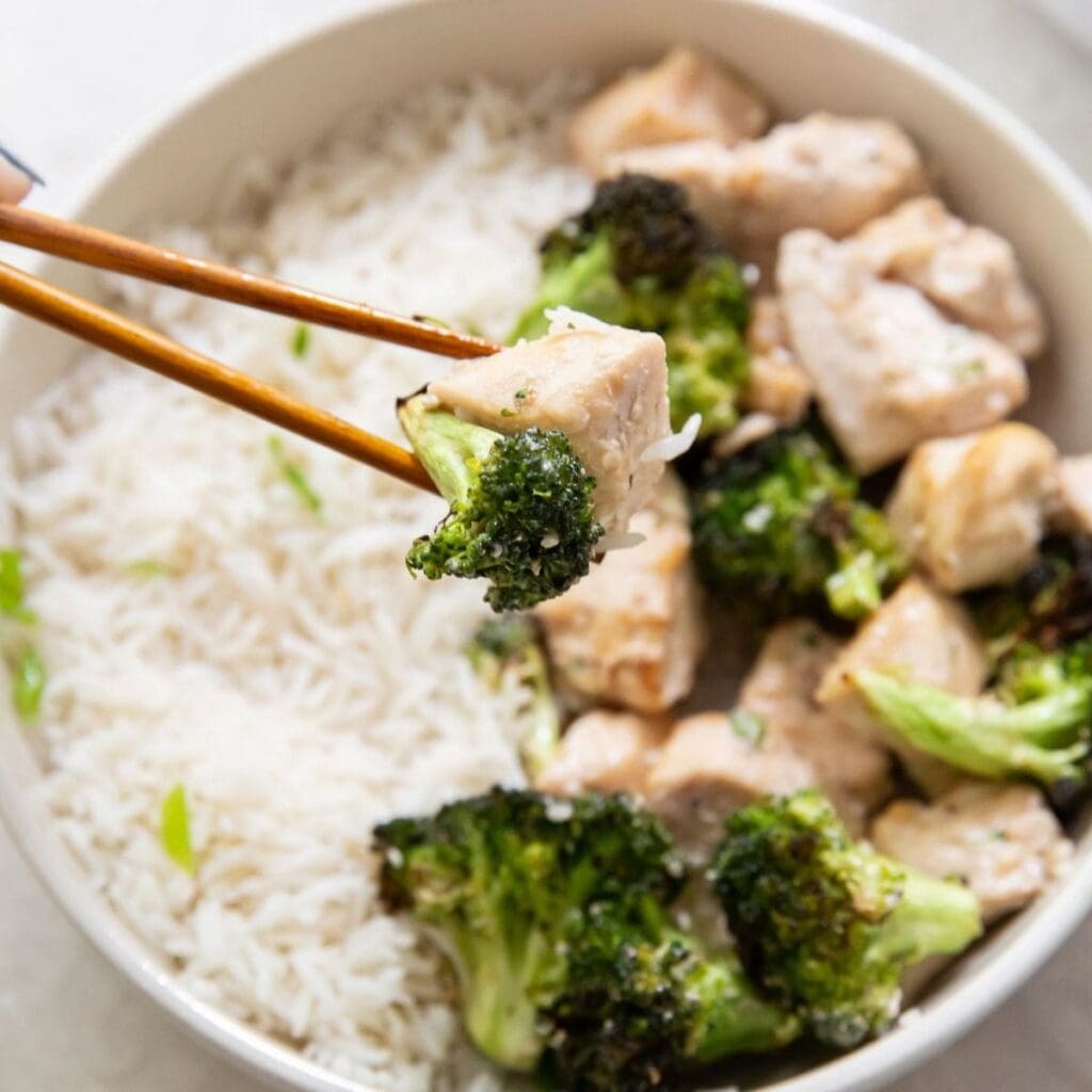 Air Fryer Chicken Recipes with Rice Close Up of a Piece of Chicken and Broccoli in Chopsticks Above a Bowl of Rice and More Chicken and Broccoli