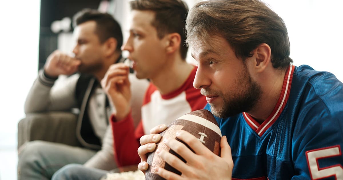 Super Bowl Air Fryer Recipes Close Up of Three Men Sitting on a Couch Wearing Sports Shirts and Holding a Football