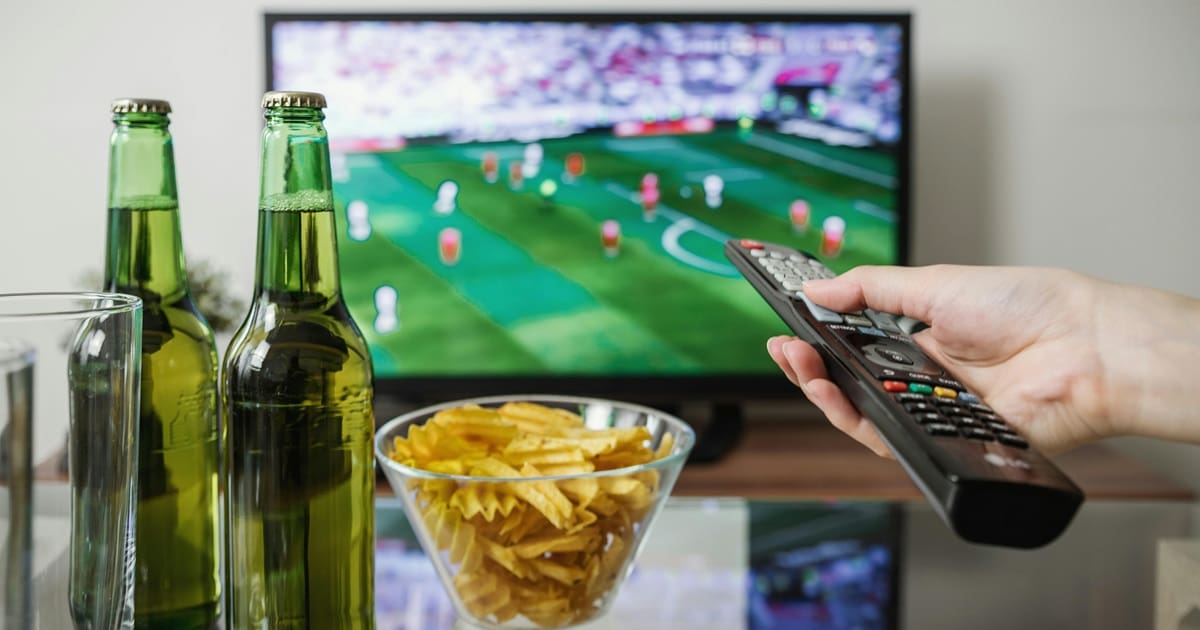 Super Bowl Air Fryer Recipes Close Up of a Person's Hand Holding a Remote Control Toward a TV with Football On