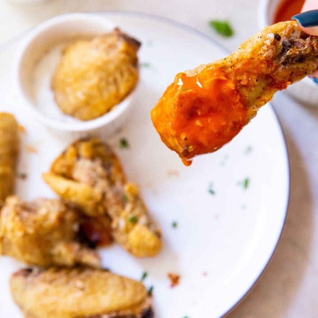 Fourth of July Air Fryer Recipes a Person Holding a Wing Dipped in Buffalo Sauce Over a Plate of Wings with a Small Ramekin of Ranch Dressing