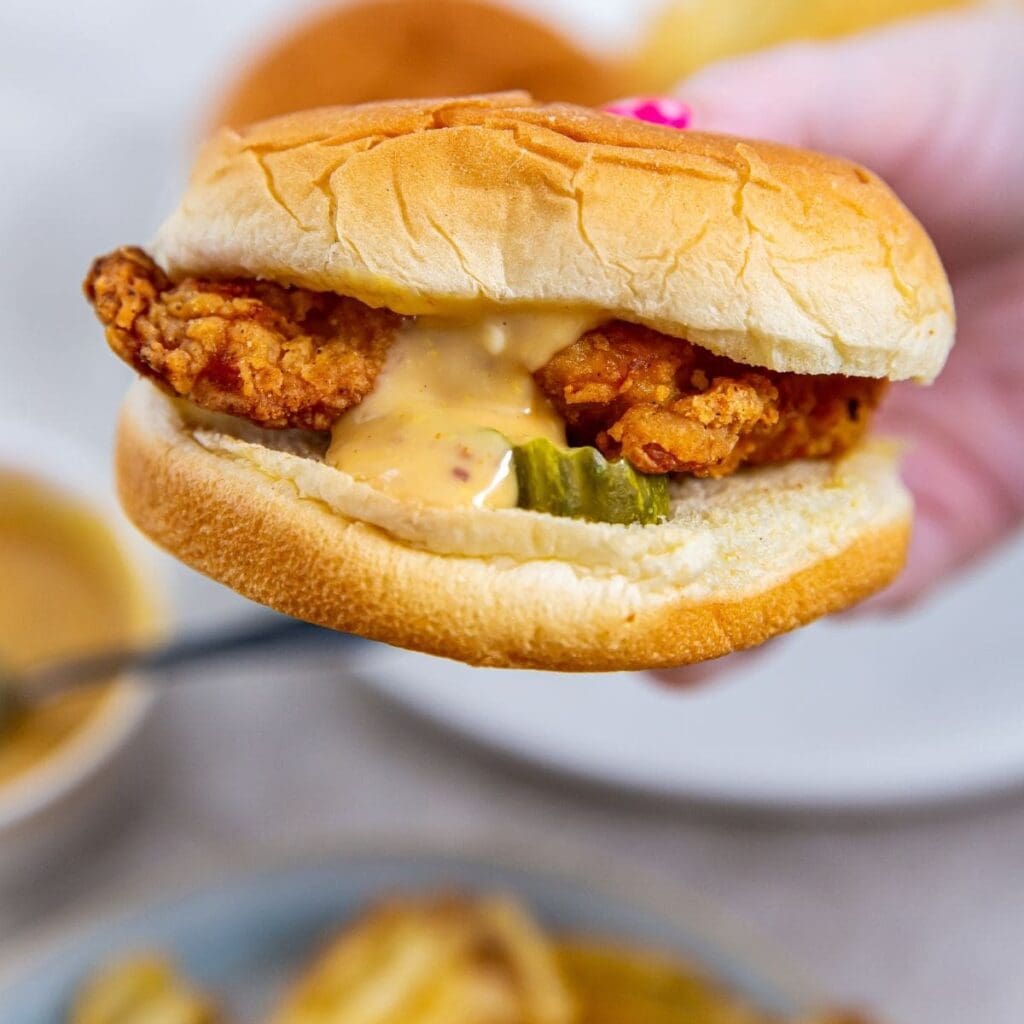 Essential Summer Air Fryer Recipes a Person's Hand Holding Up a Chicken Sandwich Dripping with Sauce Over a Plate of Fries and a Small Bowl of Sauce