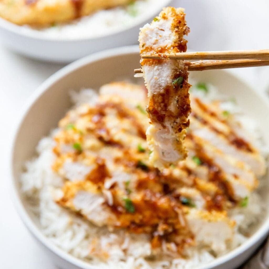 Chicken Recipes You Need to Make Close Up of piece of Katsu Chicken between Two Chopsticks Above a Bowl of Katsu Chicken with Rice